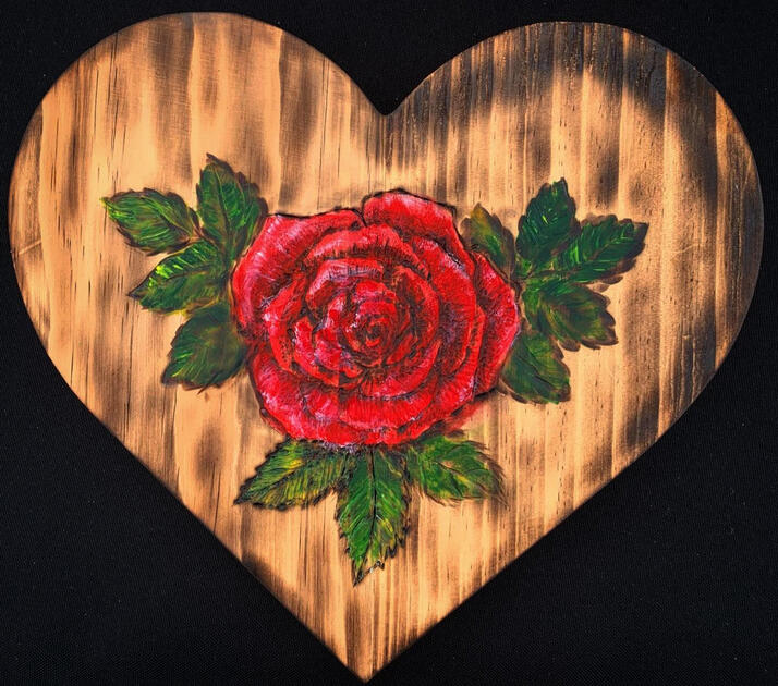 A woodburning on a heart-shaped slab of wood. It's decorated with a rose and leaves, woodburned and then painted with acrylic.